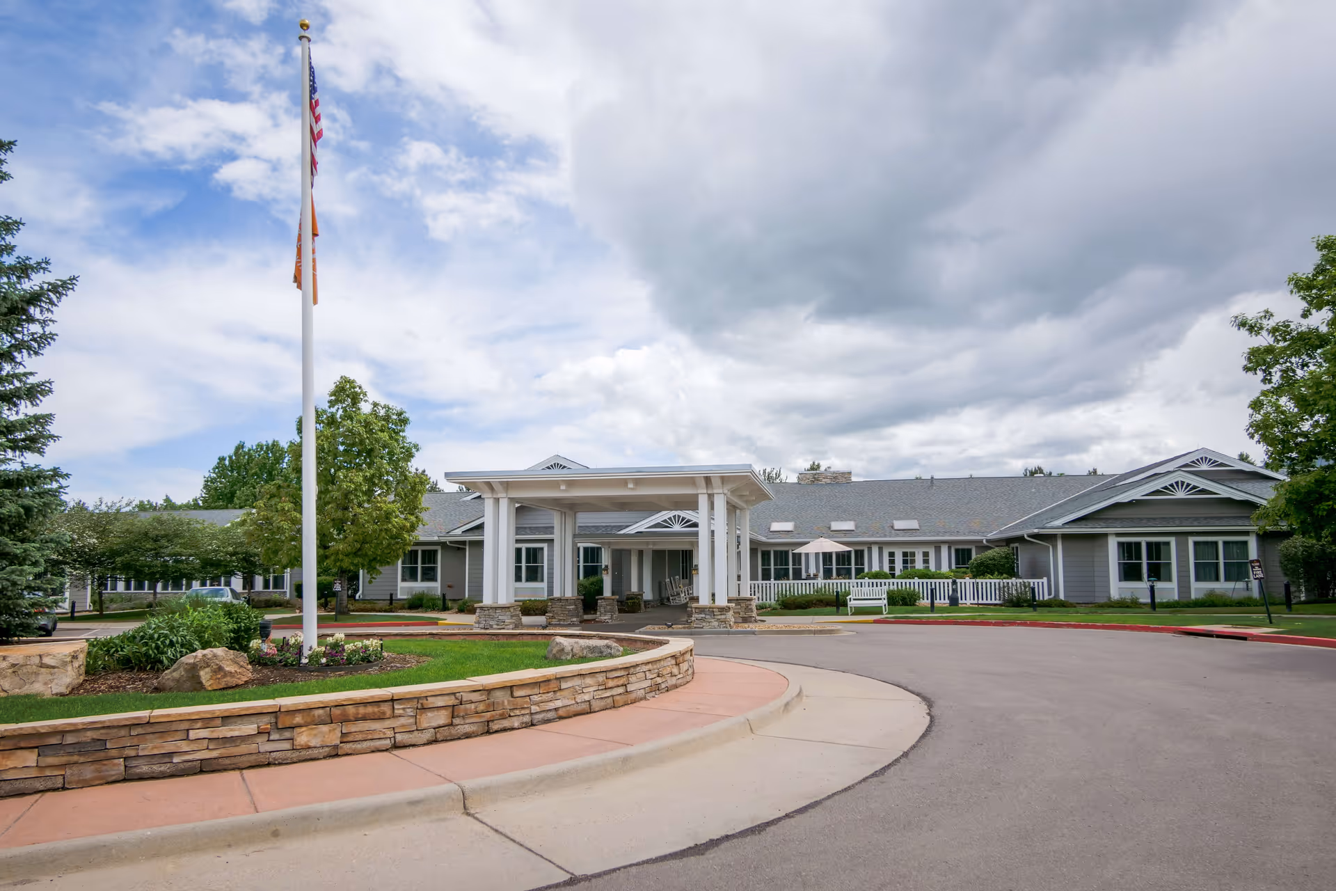 Exterior view of a single-story senior living facility building with a covered entrance, a flagpole with American and other flags, landscaped greenery, and a curved driveway under a partly cloudy sky.