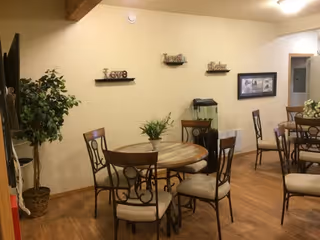 Interior view of a dining area in an assisted living facility with round wooden tables and cushioned chairs. The walls are decorated with small shelves holding decorative items and a fish tank. There is a large potted plant in the corner and framed pictures on the wall.