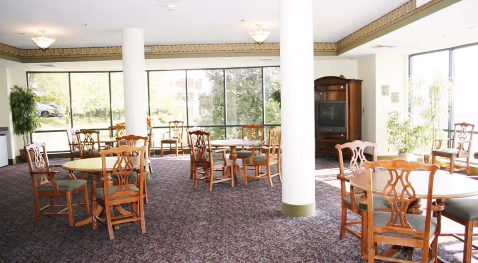 Bright communal dining room with round wooden tables and chairs, large windows, carpeted floor, and a TV cabinet.