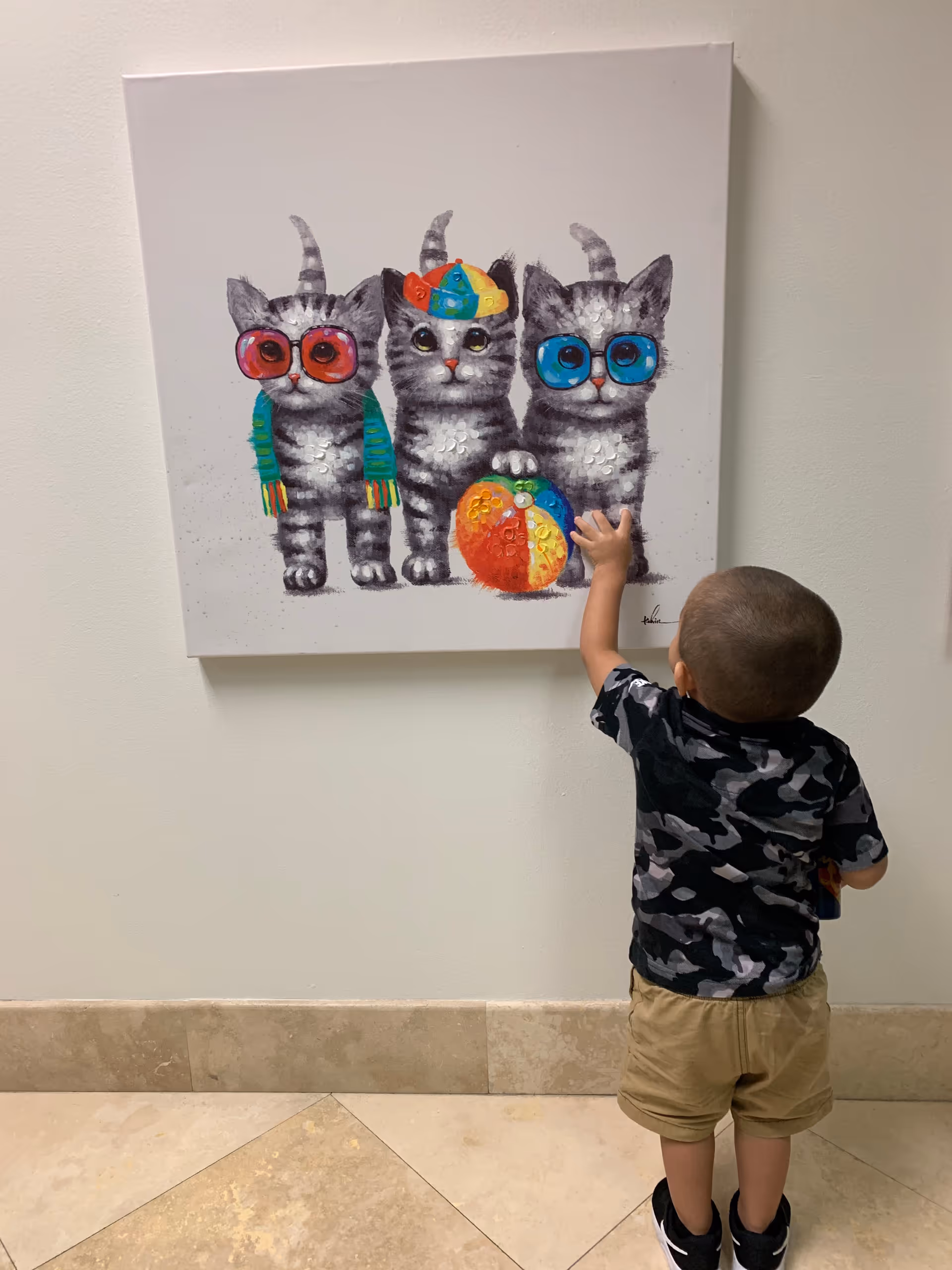 Young child touches a colorful painting of three cats wearing sunglasses on a wall.