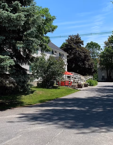 White multi-story assisted living building with a ramp and a red-umbrella seating area surrounded by trees and a driveway.