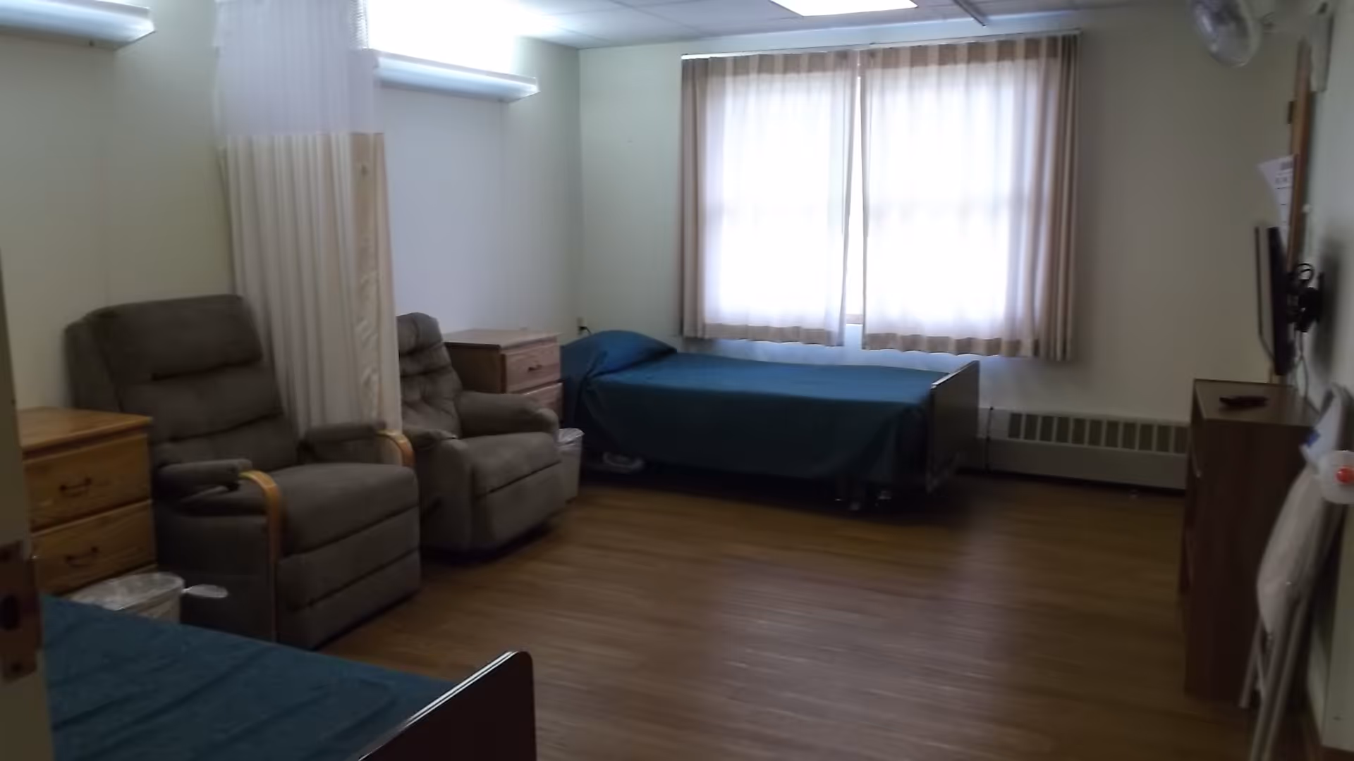A simple senior living bedroom with two single beds covered in blue bedspreads, two gray recliner chairs, wooden dressers, a window with beige curtains, and a wall-mounted TV.