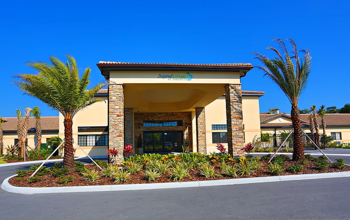 Front entrance of a Mediterranean-style senior living building with palm trees, landscaped planting beds, and a circular driveway under a clear blue sky.
