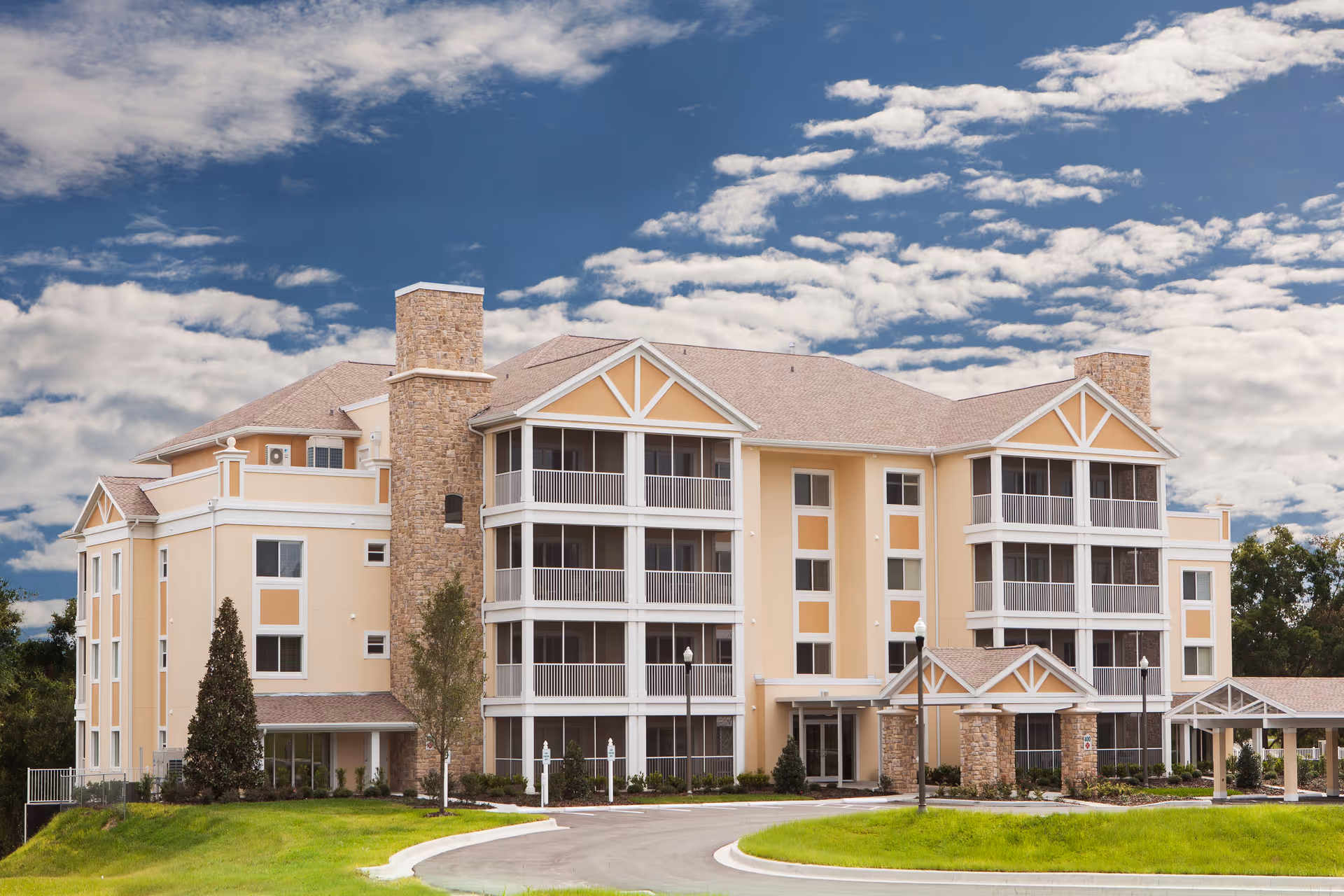 Exterior view of a multi-story retirement community building with beige walls, stone accents, screened balconies, and a covered entrance. The building is surrounded by green grass and trees under a partly cloudy blue sky.