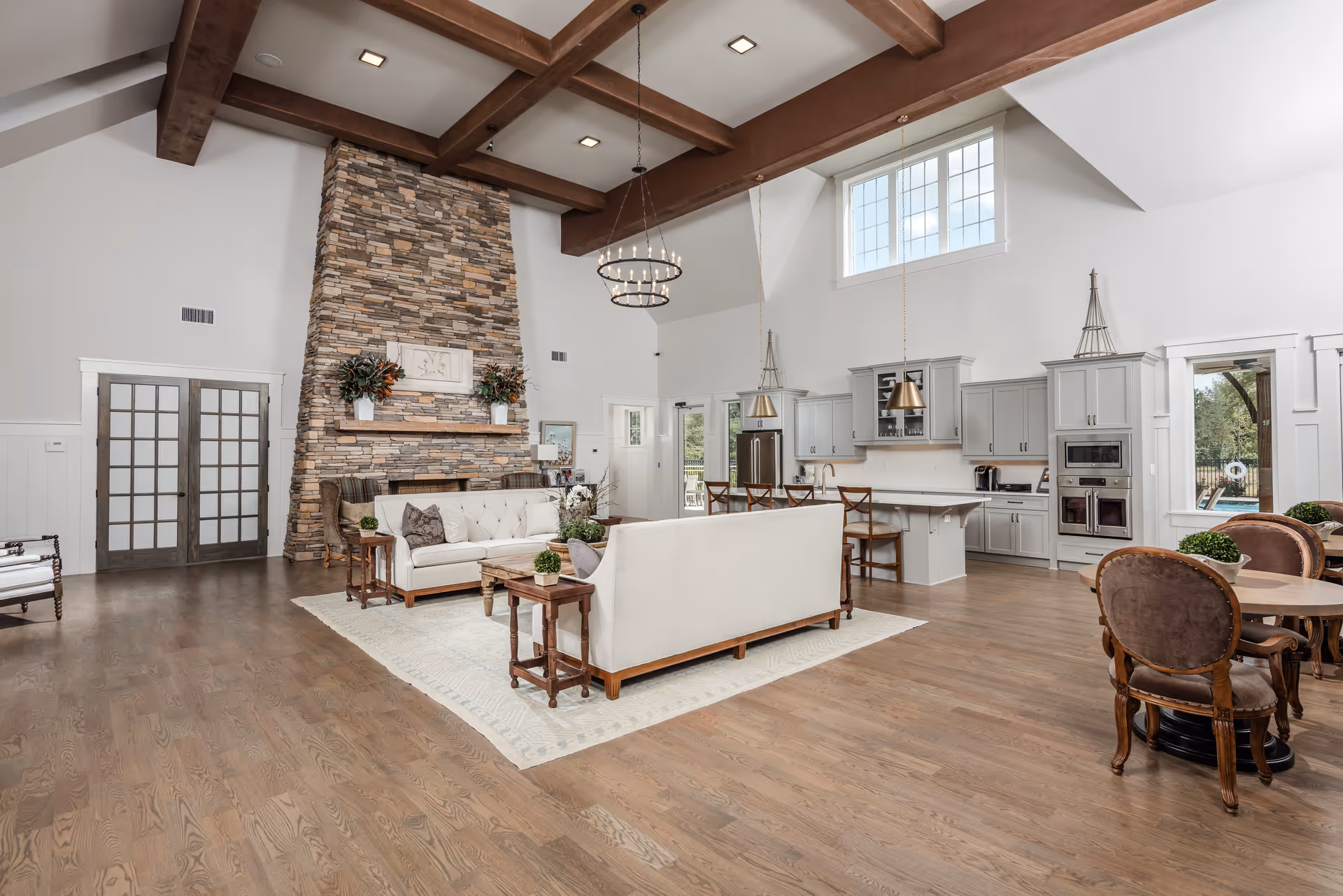 Spacious open-concept living area with high vaulted ceilings and exposed wooden beams. The room features a large stone fireplace with decorative plants on the mantel, two white sofas arranged around a wooden coffee table on a light-colored rug, and a kitchen area with white cabinetry, stainless steel appliances, and a breakfast bar with stools. There is also a round dining table with upholstered chairs near windows that let in natural light.