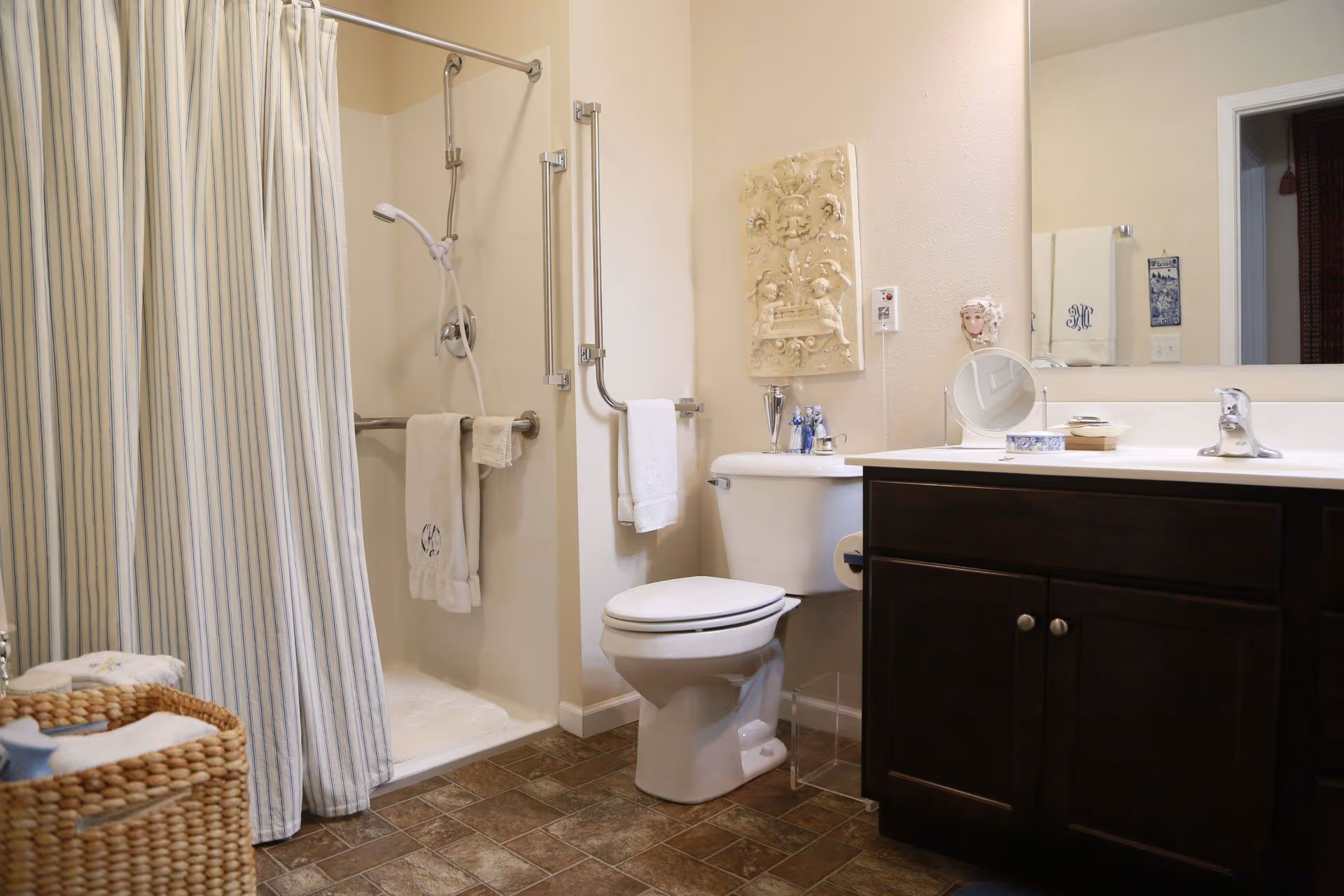 Clean accessible bathroom with shower and striped curtain, toilet, dark wood vanity with sink and mirror, and a wicker laundry basket.