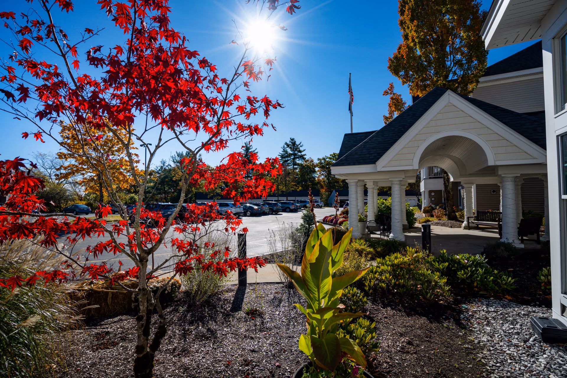 Sunlit entrance of RiverWoods Manchester with white arches, pillars, and red autumn foliage.