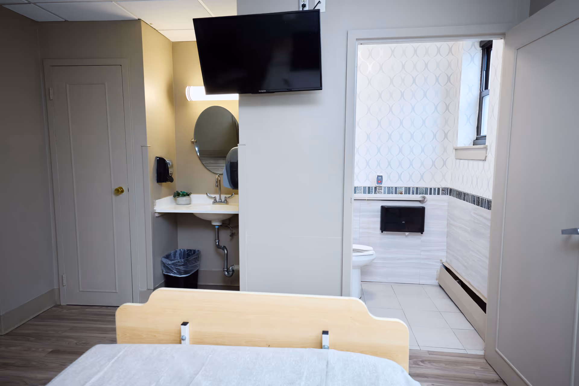Hospital-style patient room with bed, sink area, and adjoining tiled bathroom.