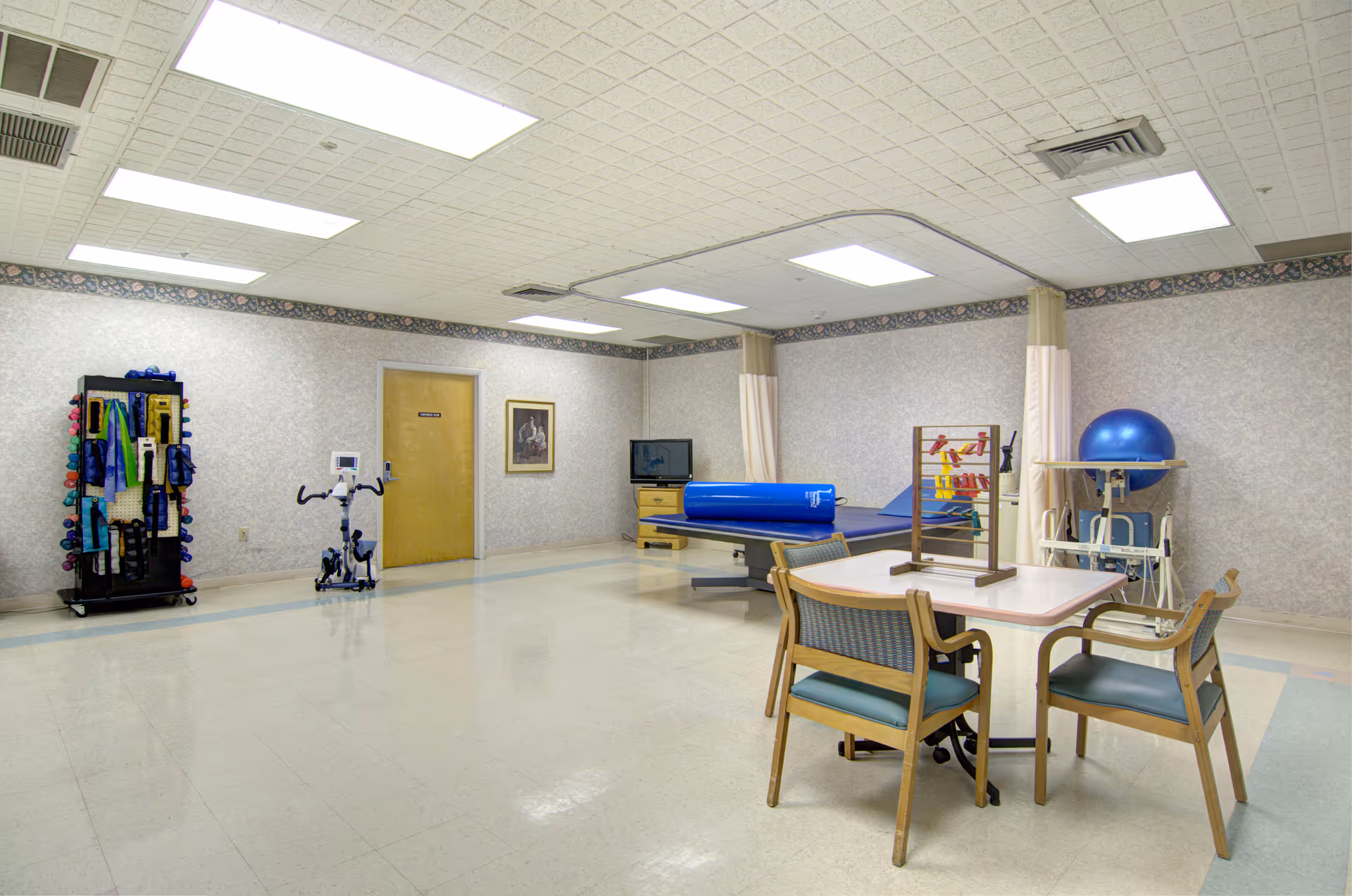 An interior recreation room in a senior facility with exercise equipment, a table, and chairs.