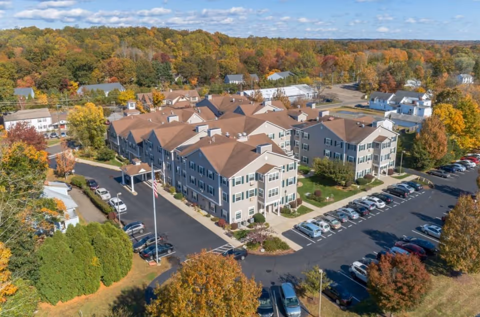 Aerial view of a senior living facility named HarborChase of Madison surrounded by trees with autumn foliage. The building is a large, multi-story structure with a brown roof and beige exterior walls. There is a parking lot with many cars parked around the building and a flagpole with an American flag near the entrance.