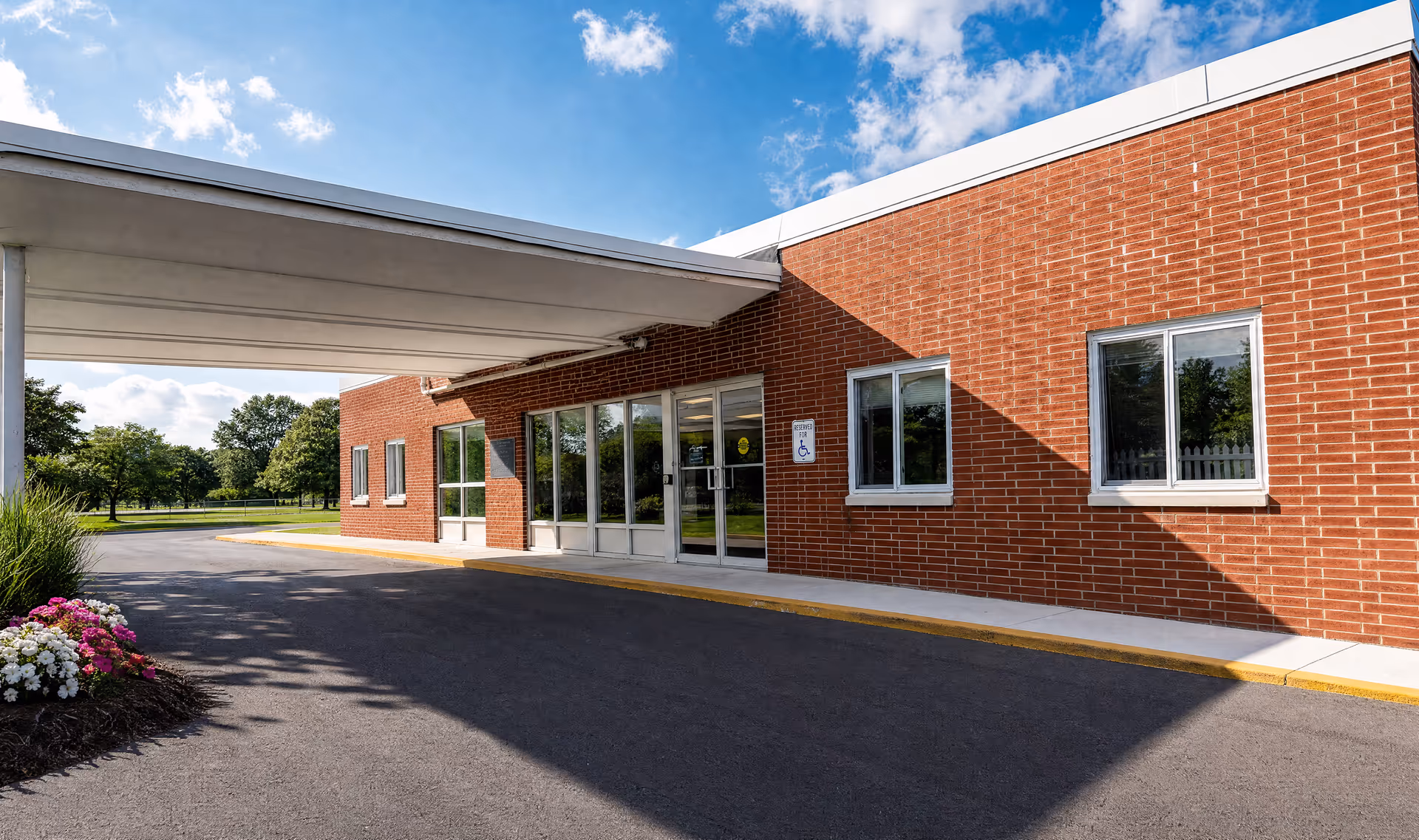 Brick assisted living facility entrance with glass doors under a covered canopy on a sunny day.