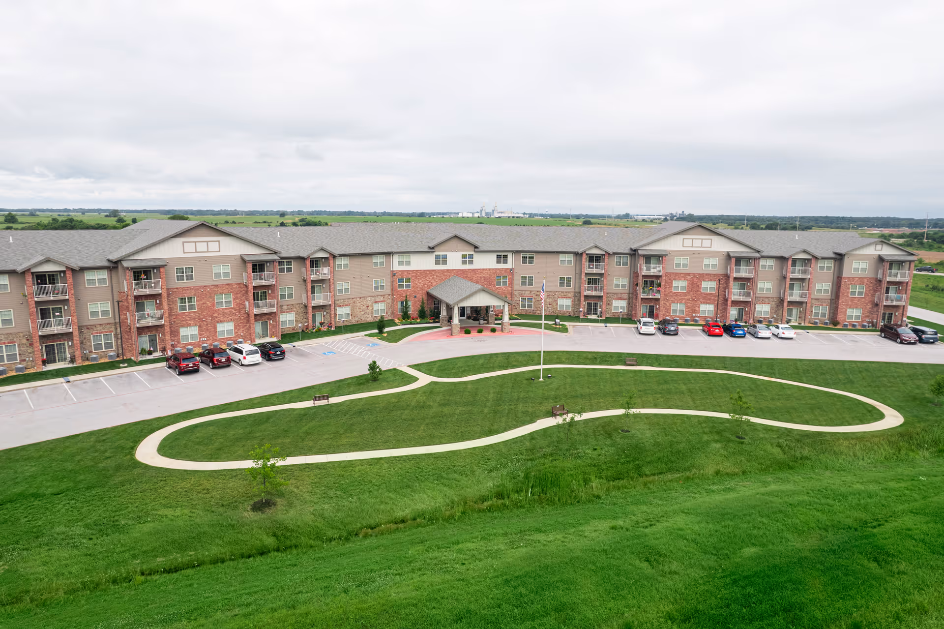 Wide exterior view of a large, three-story senior living facility building with a combination of brick and siding facade. The building has multiple balconies and windows, a covered entrance, a parking lot with several cars, and a curved walking path on a well-maintained green lawn in front.