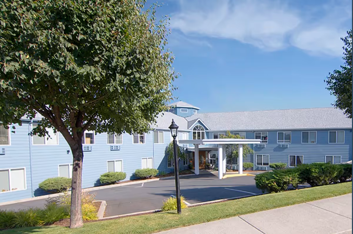 Blue two-story senior living building with a covered entrance and surrounding landscaping.