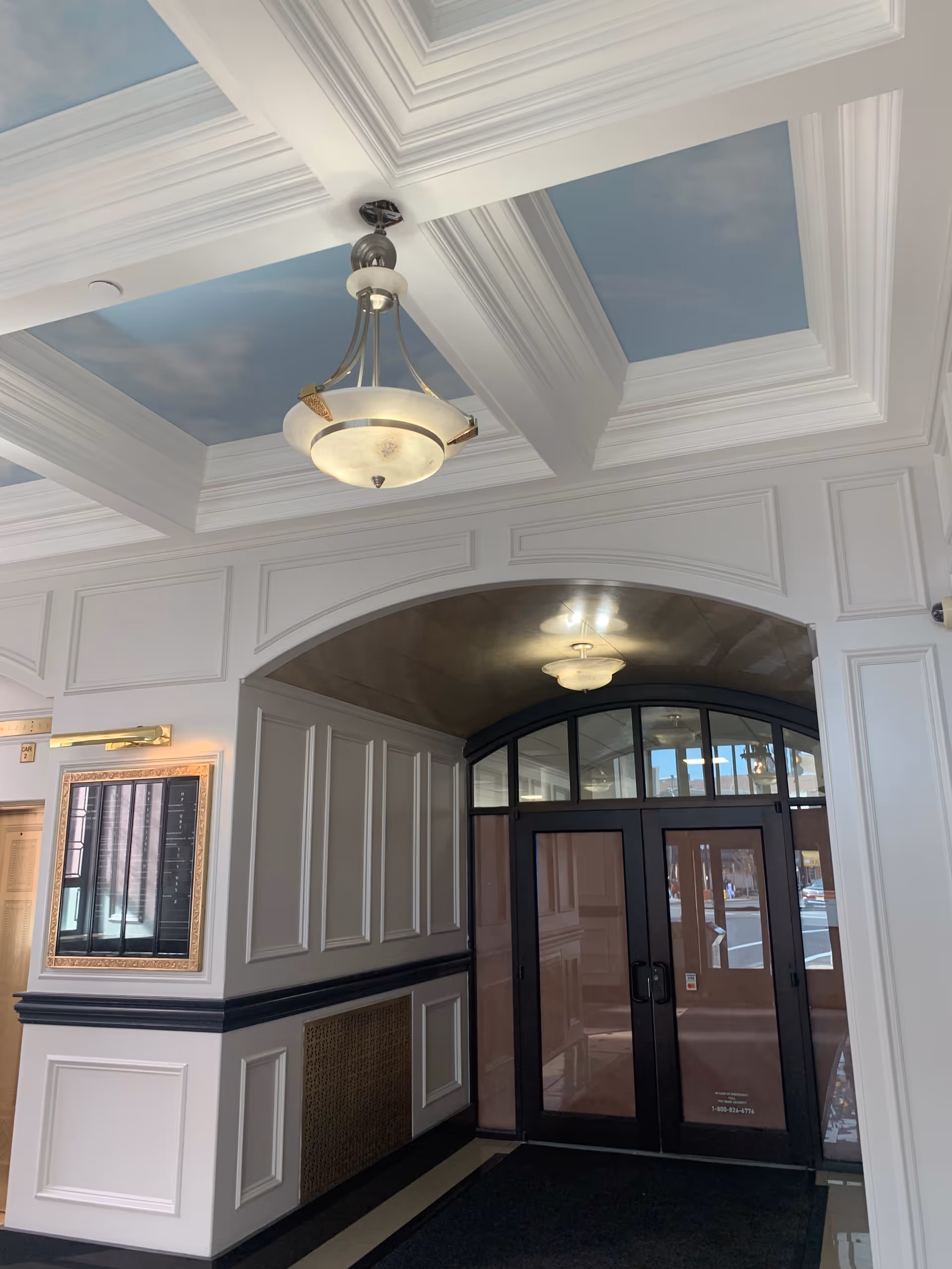 Bright interior lobby with paneled walls, arched doorway, and decorative ceiling chandeliers.
