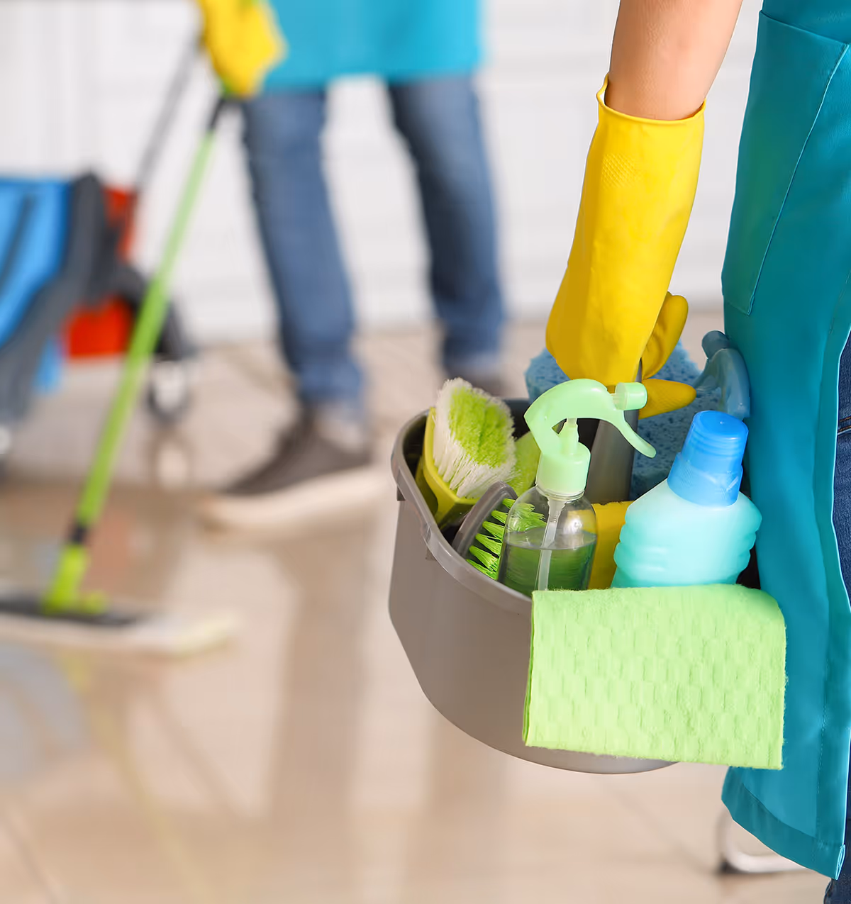 Cleaning supplies bucket and gloves held by a janitor in a bright interior hallway.