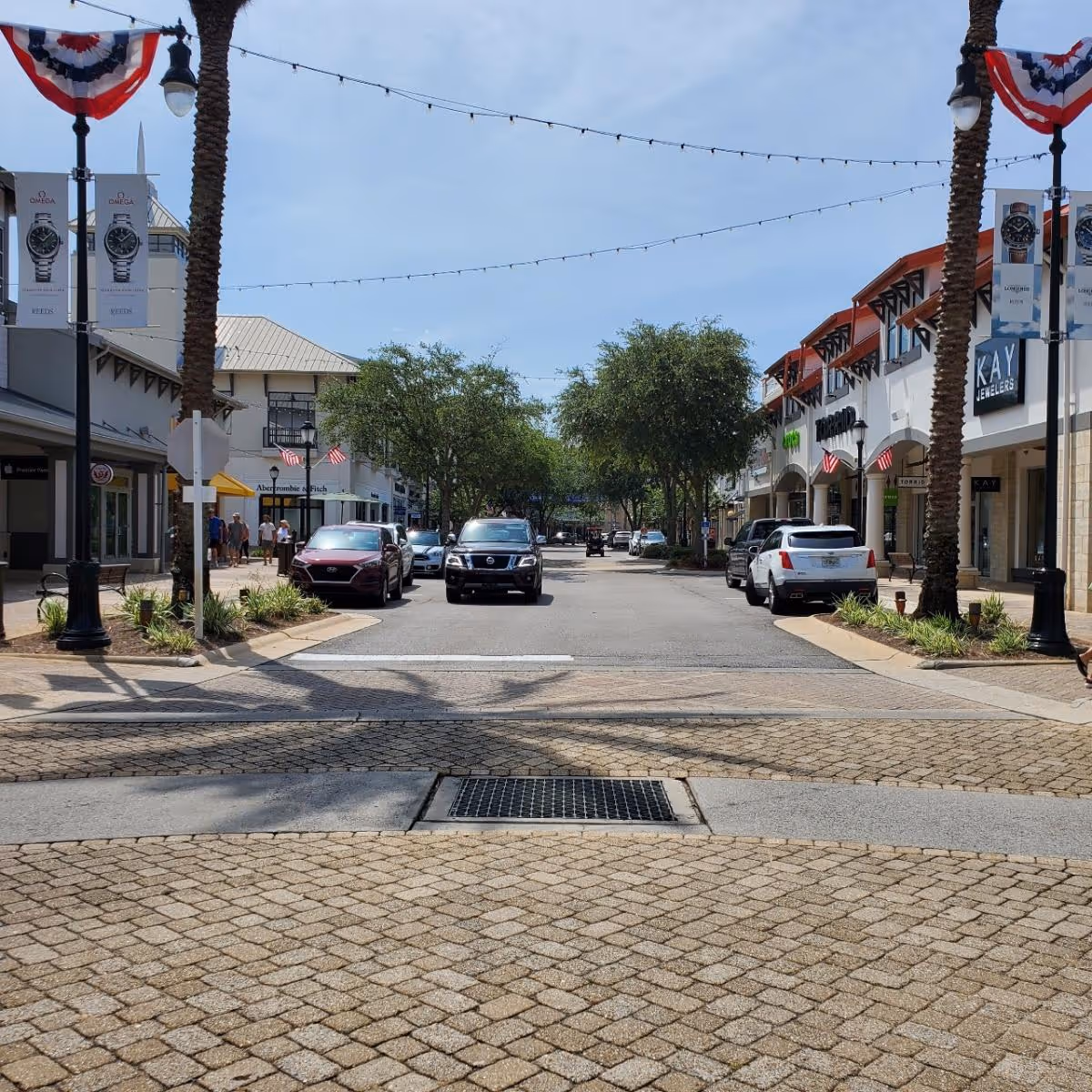 Sunny outdoor shopping street with storefronts, bunting, and parked cars.