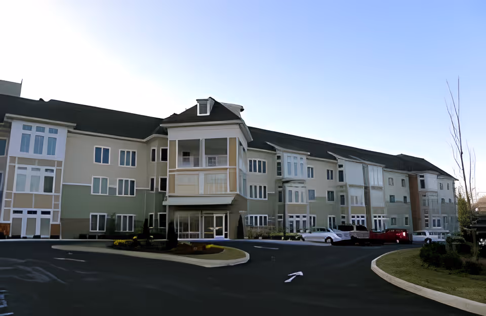 Exterior view of a large, multi-story senior living facility building with multiple windows and a driveway in front. Several cars are parked near the entrance, and there is a clear blue sky above.