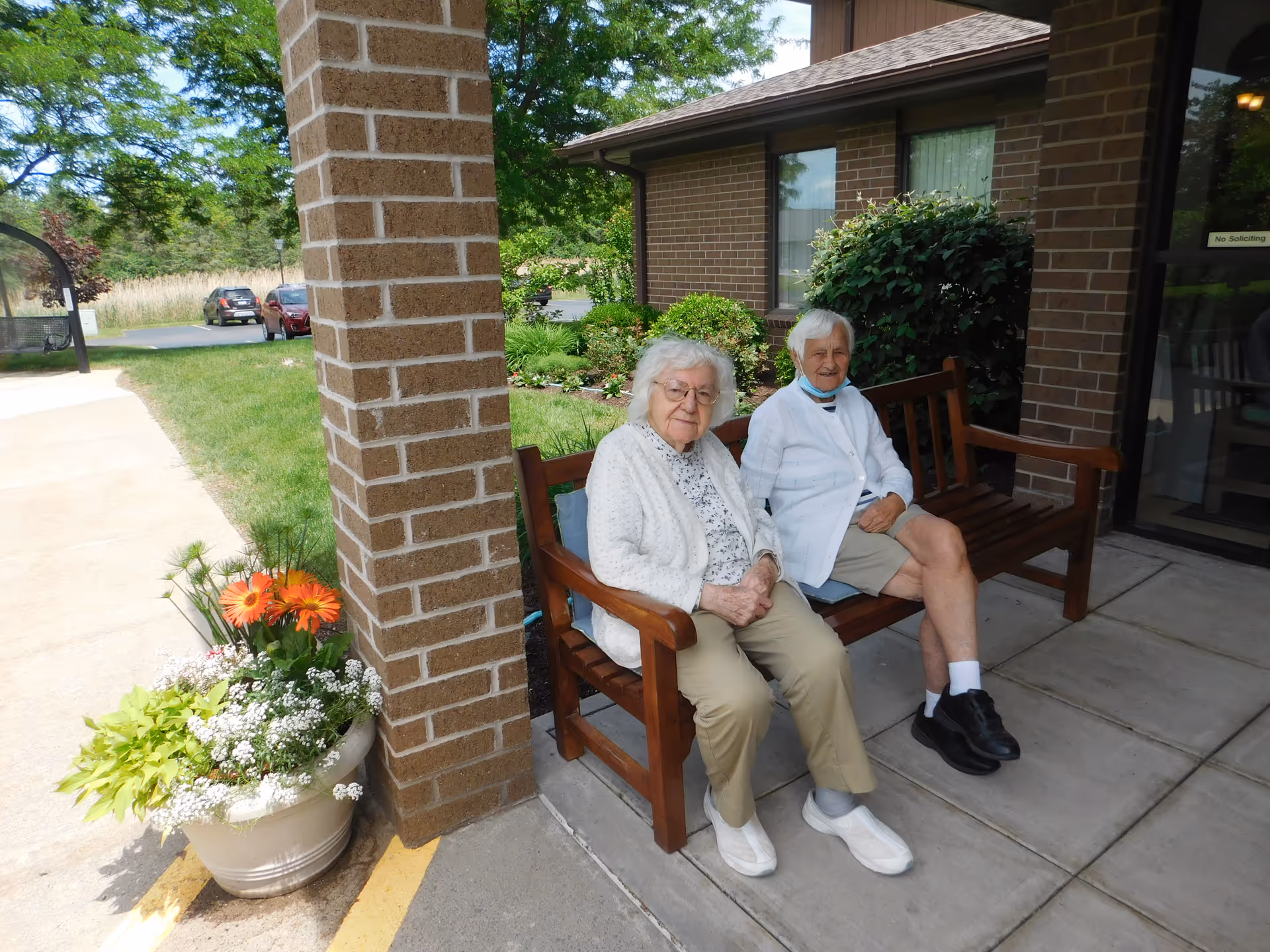 Two elderly women sit on a wooden bench outside a brick building with potted flowers.