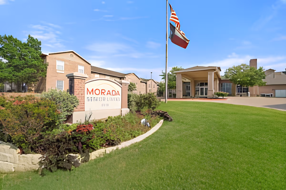 Front entrance of the Morada Pantego senior living facility with a sign, flagpoles, landscaped lawn, and building facade.