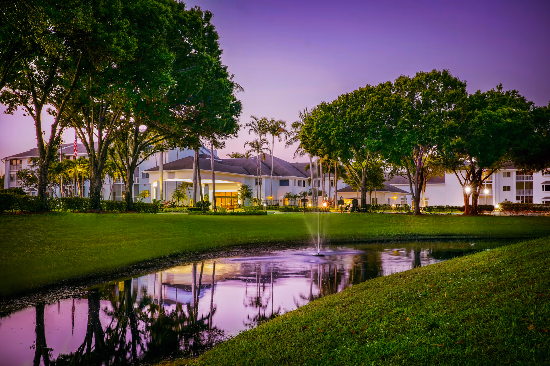 Evening view of Vi at Lakeside Village with a lit entrance, trees, and a reflective pond.