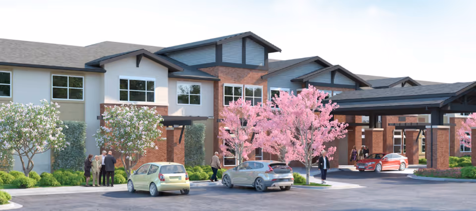 Front exterior of a modern senior living building with a covered porte-cochère, parked cars, and pink flowering trees.