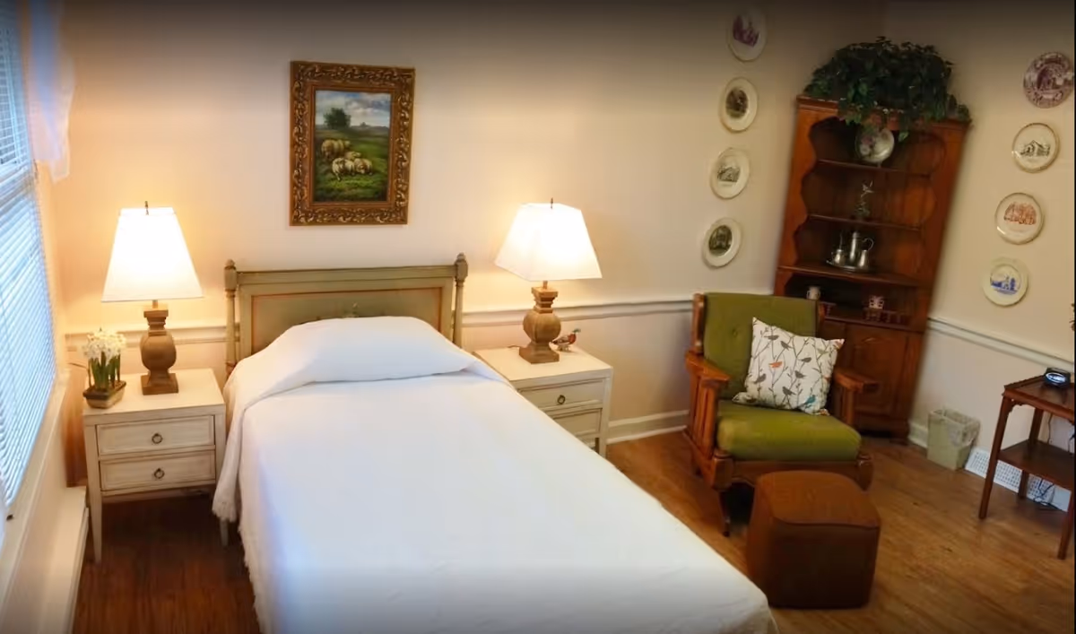 Neatly arranged bedroom with a single bed flanked by nightstands and lamps, an armchair, and shelving with decorative plates.