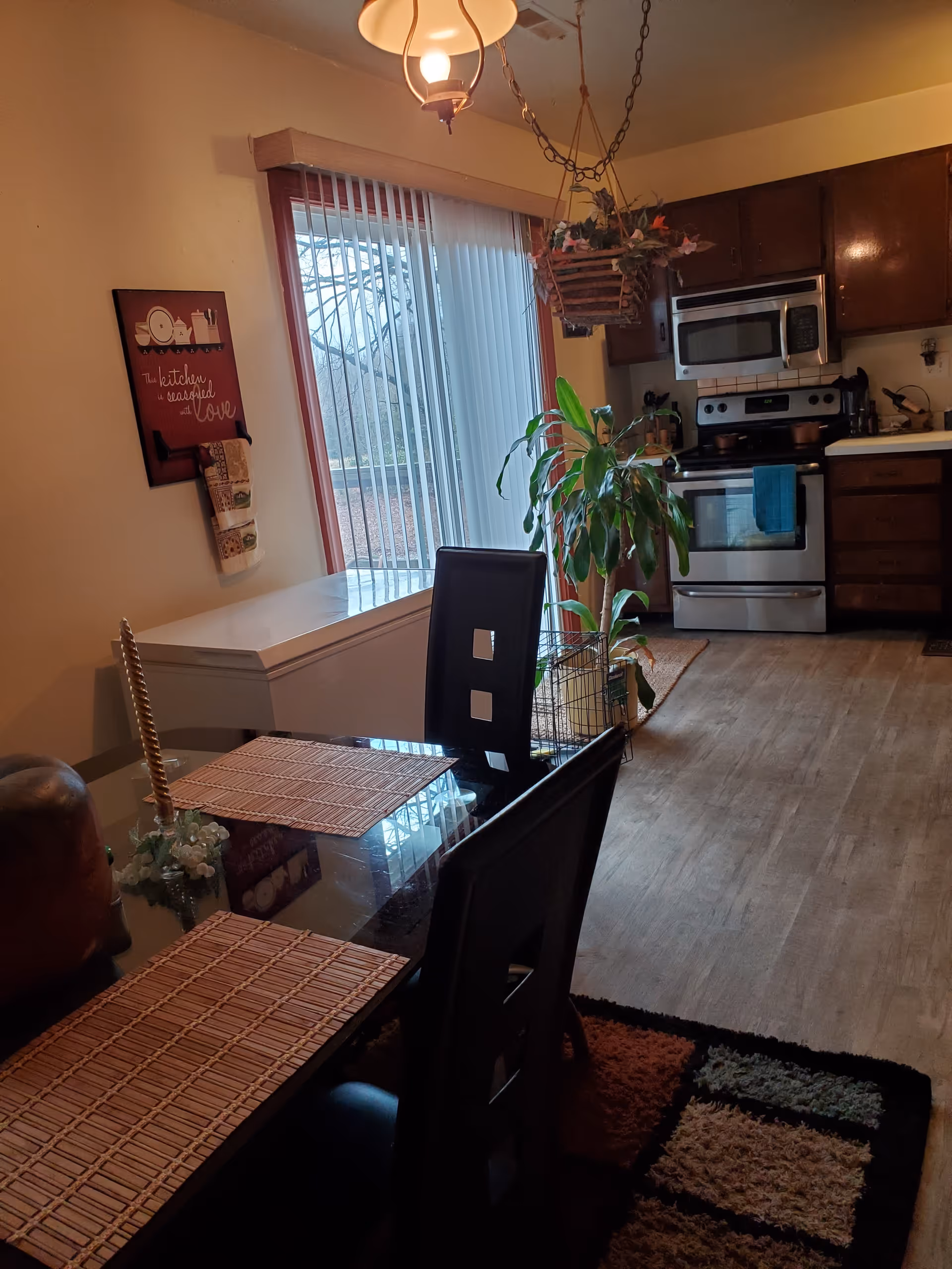 Dining area with glass table, black chairs, window blinds, and a kitchen in the background.