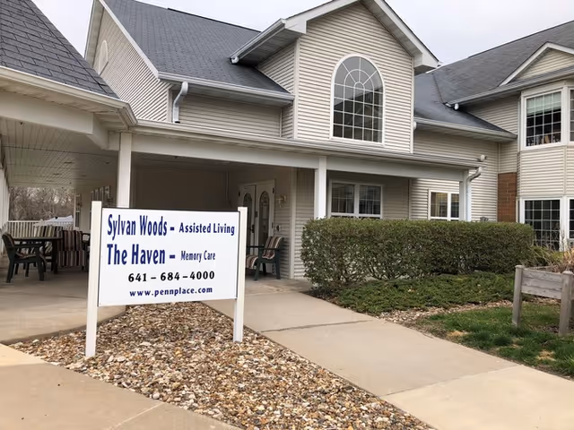 Exterior view of a senior living facility building with beige siding and a gray roof. There is a covered entrance with chairs and a sign in front that reads 'Sylvan Woods - Assisted Living, The Haven - Memory Care, 641-684-4000, www.pennplace.com'. The area around the entrance has landscaping with bushes and rocks.