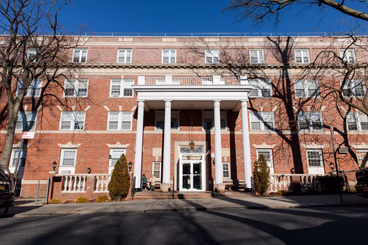 Front facade of a red-brick, multi-story residential building with white entrance columns and leafless trees casting shadows.