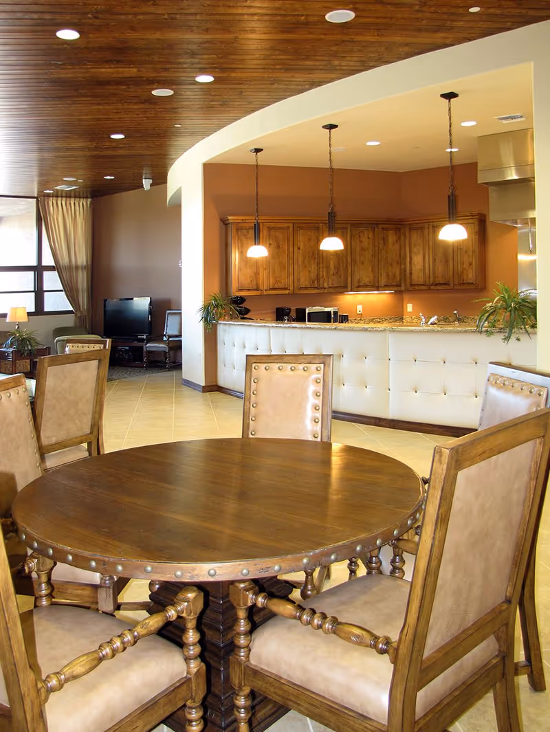 Communal dining area with a round wooden table and chairs facing a bar-style kitchen with pendant lights and wood cabinets.
