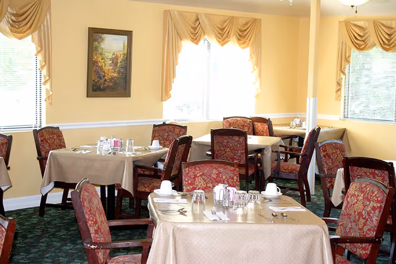 Bright dining room with multiple tables set with tablecloths and patterned chairs beneath windows with gold valances.