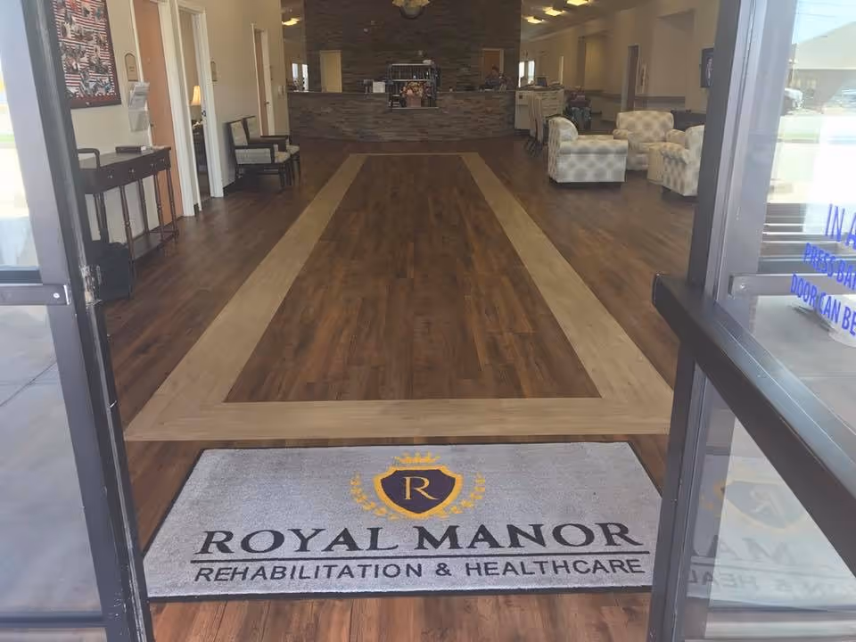 View through open glass doors into the entrance lobby of Royal Manor Rehabilitation and Healthcare, showing a floor mat with the facility's logo and name, wood-patterned flooring, a reception desk in the background, and seating areas with armchairs on the right side.