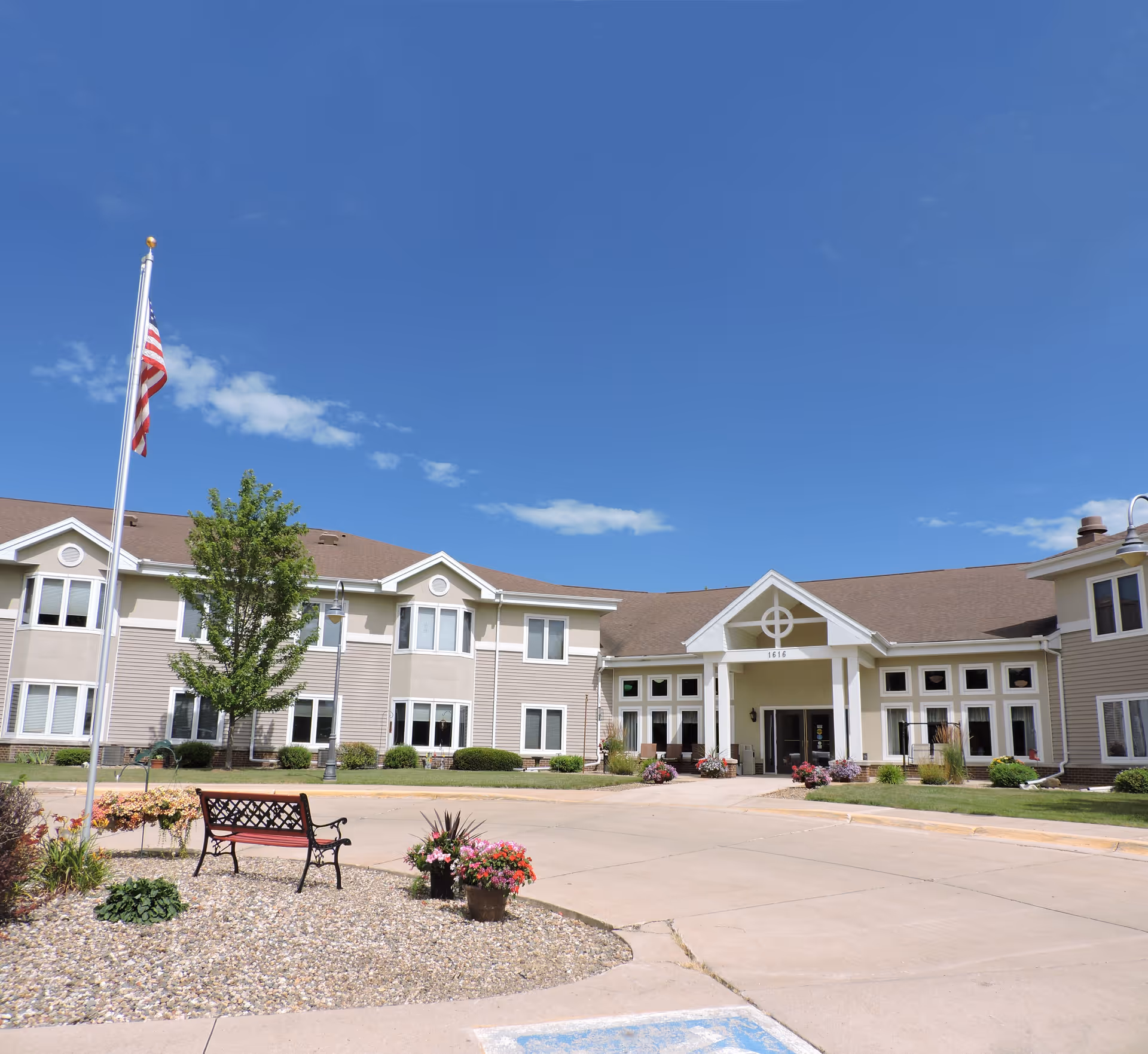 Front exterior of a two-story senior living facility with an entrance portico, American flag, benches, and landscaped planters under a clear blue sky.