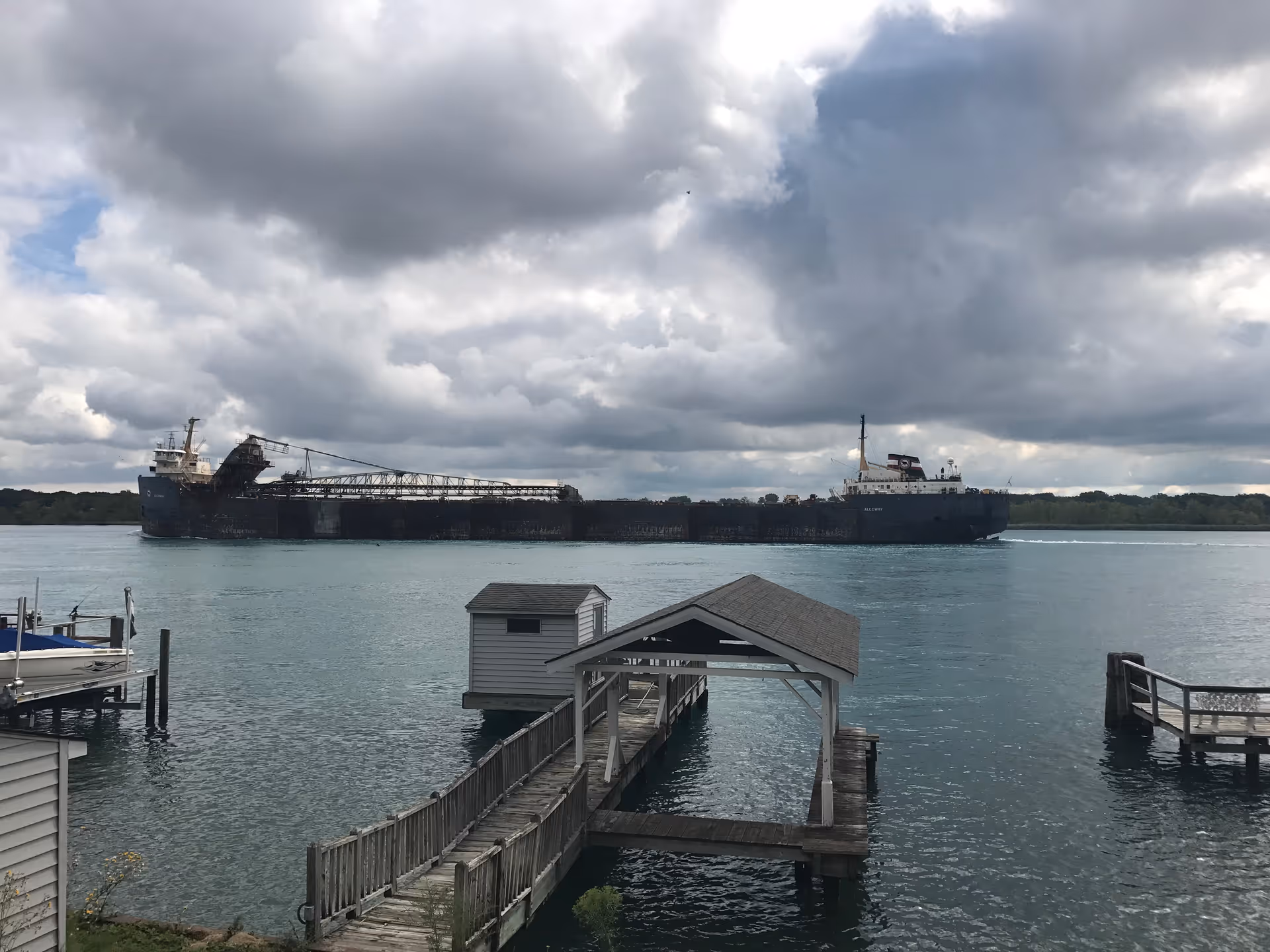A large cargo ship sails past a wooden dock along calm water under cloudy skies.
