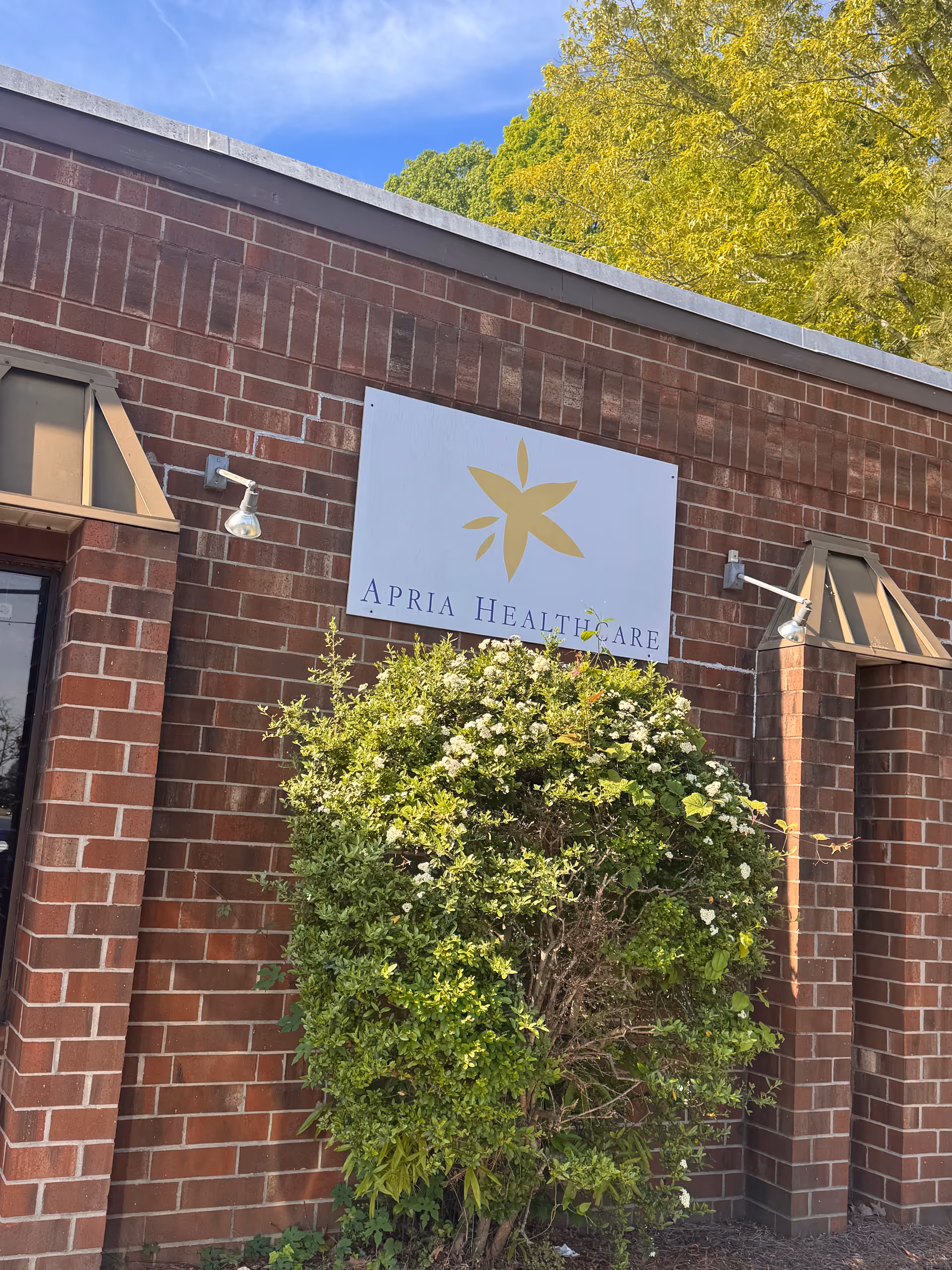 Brick building exterior with Apria Healthcare sign and front greenery.