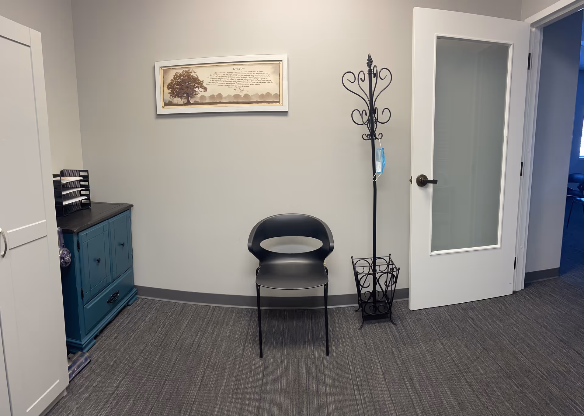 Small waiting area in senior facility: chair, coat rack, cabinet, wall art, glass door.