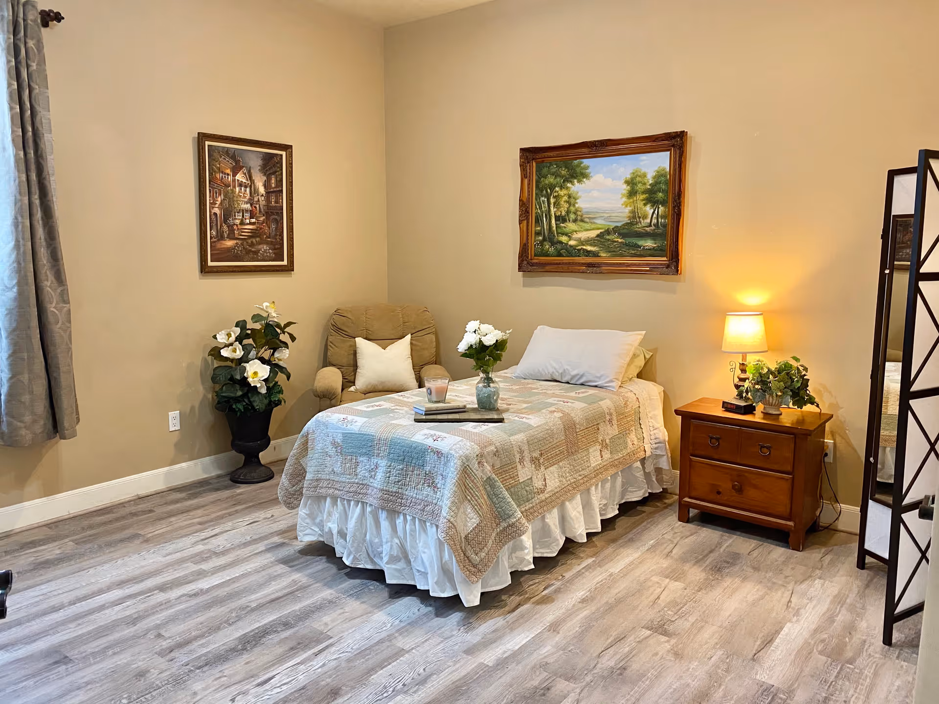 A cozy bedroom with a single bed covered in a patchwork quilt and white bed skirt. A beige armchair with a white pillow is placed next to the bed. On the right side of the bed is a wooden nightstand with a lit lamp and a small plant. Two framed paintings hang on the beige walls, one depicting a village scene and the other a landscape with trees and a path. A tall potted plant with white flowers is near the armchair, and a folding screen with a mirror is partially visible on the right. The floor is light-colored wood, and a window with gray curtains is on the left.