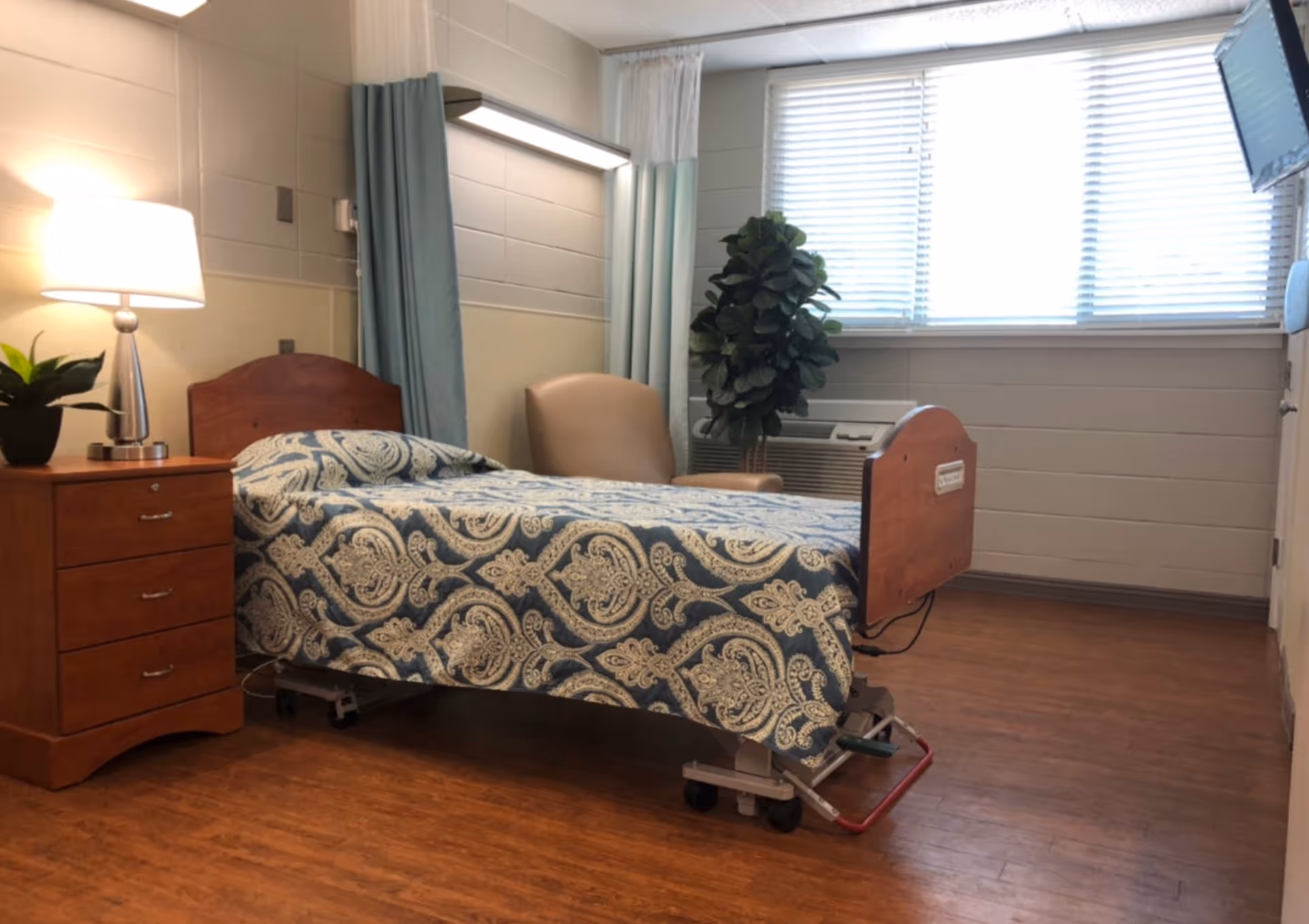 Private senior living facility bedroom with a wooden dresser, patterned bed, chair, curtains, and plant.