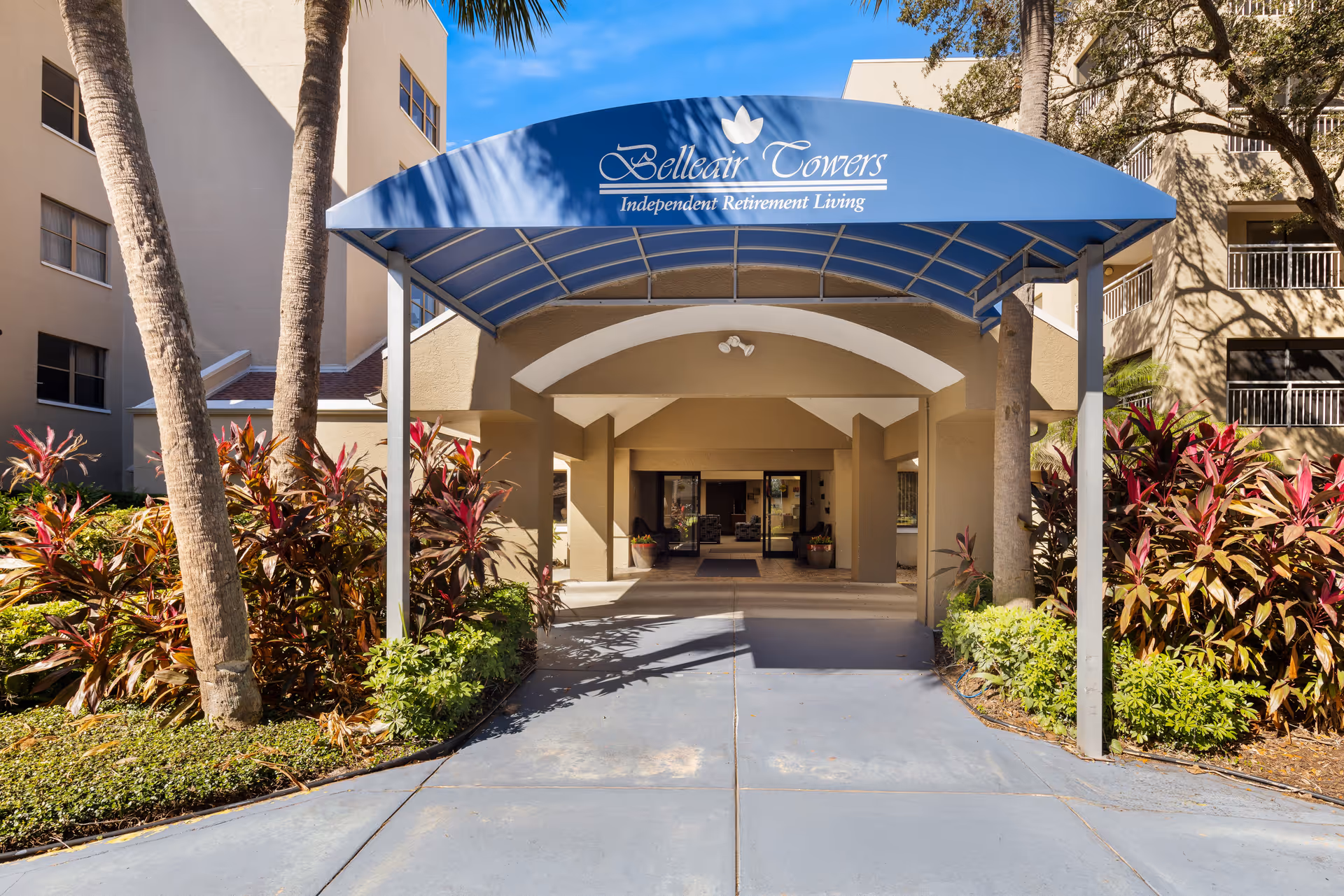 Exterior entrance of Belleair Towers with a blue canopy, palm trees, and tropical landscaping.