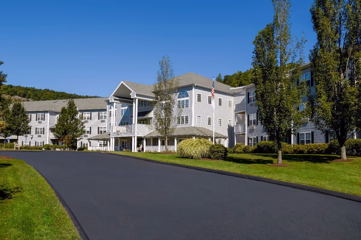 Front exterior of a multi-story senior living building with a paved driveway, green lawns and trees under a clear blue sky.