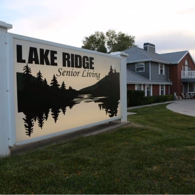 A large outdoor sign for Lake Ridge Senior Living featuring a silhouette of trees and a lake, with a two-story residential building in the background surrounded by grass and a paved walkway.