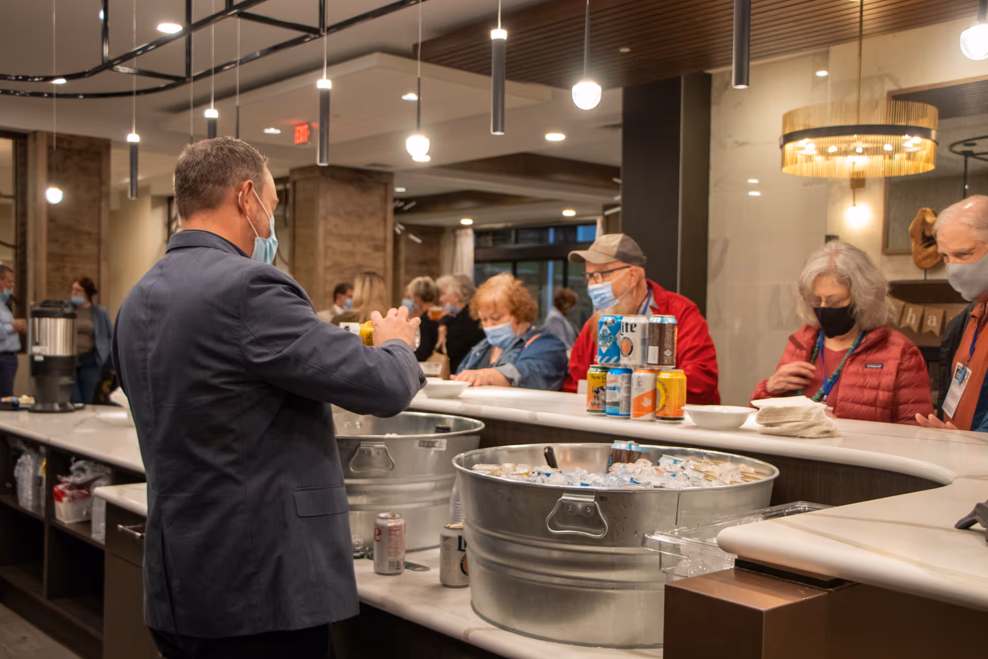 A man wearing a mask and a suit jacket is serving canned beverages from large metal tubs filled with ice to a group of elderly people who are also wearing masks and standing at a counter in a well-lit indoor setting.