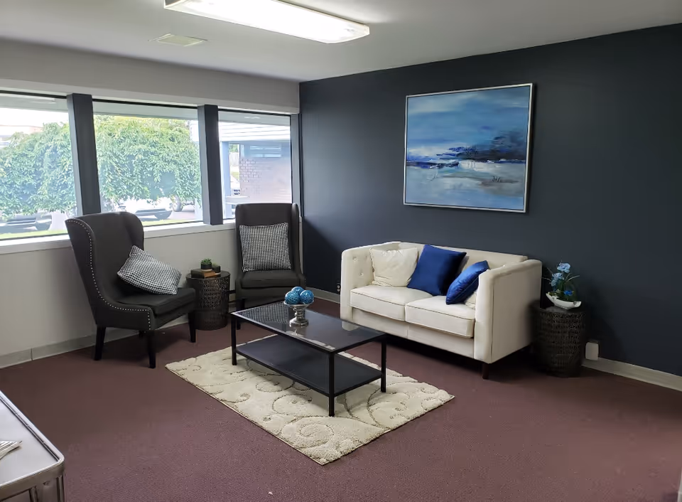 Light-filled senior living lounge with a white sofa, dark chairs, coffee table, rug, and art.