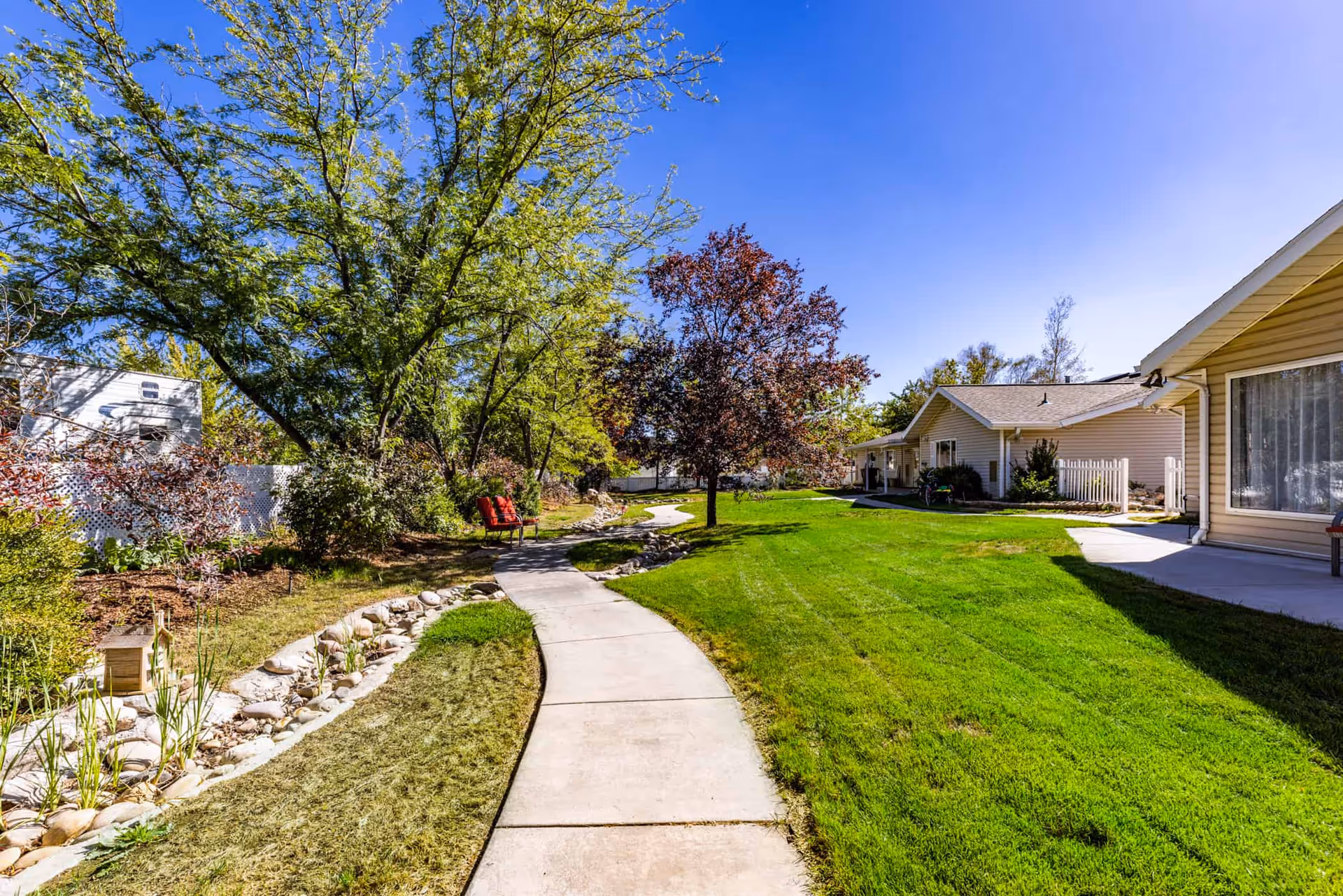 A sunny outdoor pathway winding through a green lawn with trees and bushes on the left side and single-story beige buildings on the right side under a clear blue sky.
