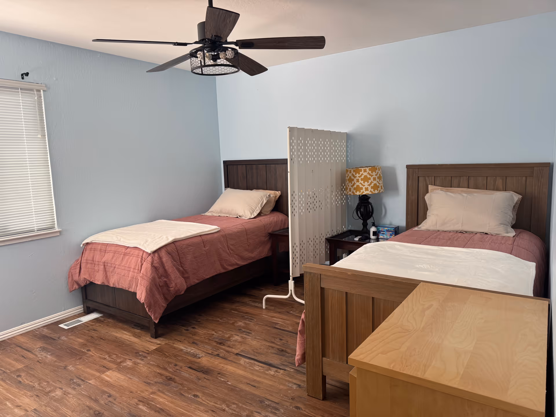 A bedroom with two single beds separated by a white folding privacy screen. Each bed has a pillow and a blanket, with a nightstand between them holding a lamp with a yellow patterned shade and some small items. The room has light blue walls, a window with blinds, a ceiling fan with dark blades, and wooden flooring.
