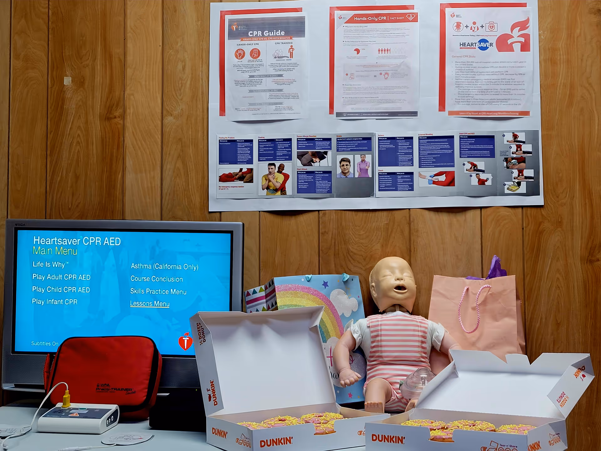 Interior room with CPR posters, a monitor, a baby doll, and pizza boxes on a table.