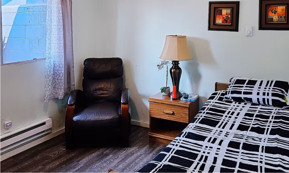A bedroom interior with a single bed, armchair, nightstand, lamp, and window.