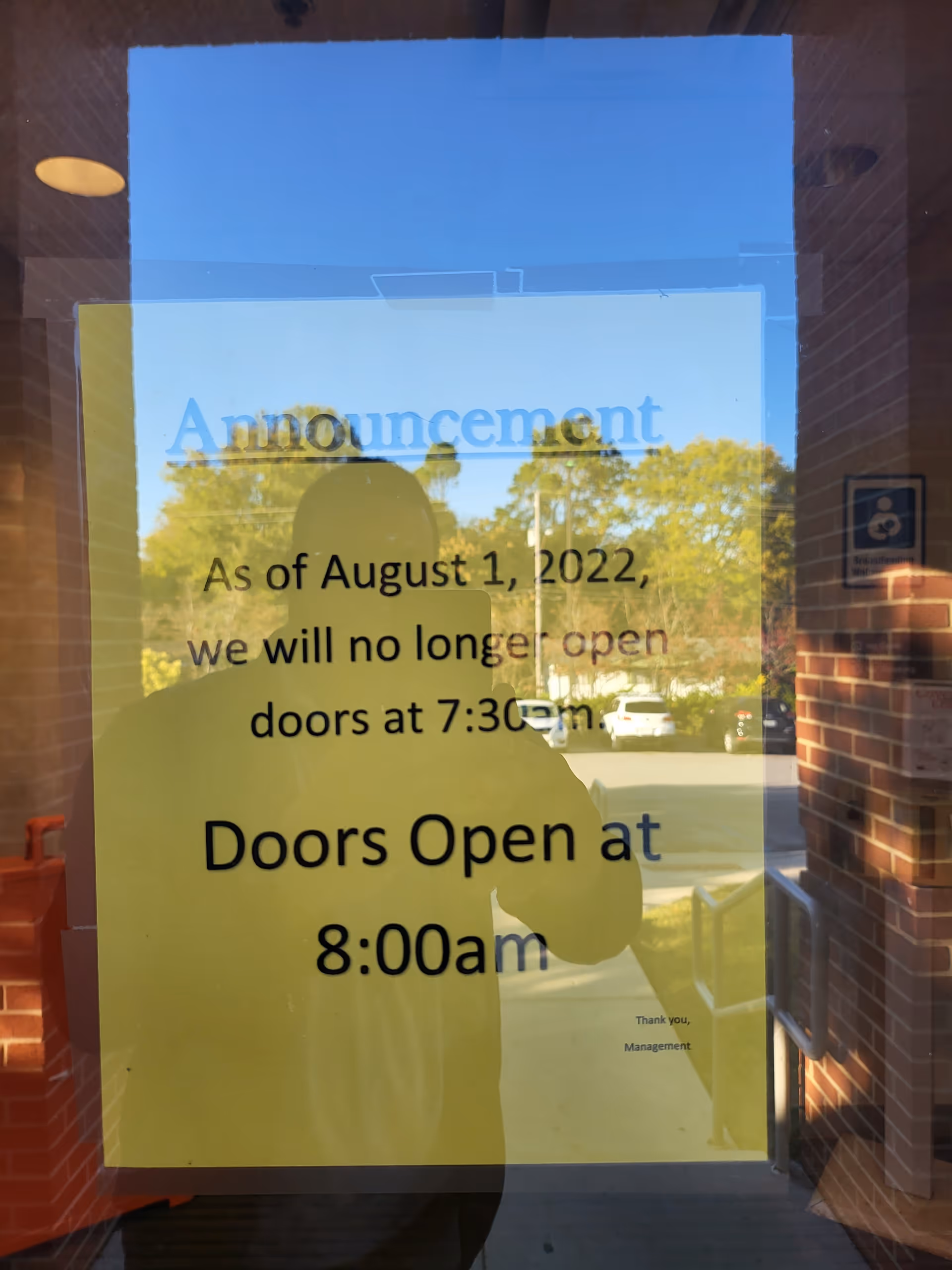 Yellow announcement sign on glass door, reflecting trees and a person silhouette.