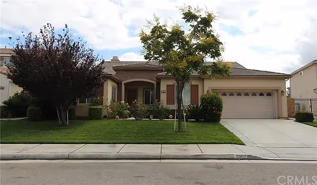Single-story residential house with a two-car garage, a small front porch, and a well-maintained lawn with trees and shrubs under a partly cloudy sky.
