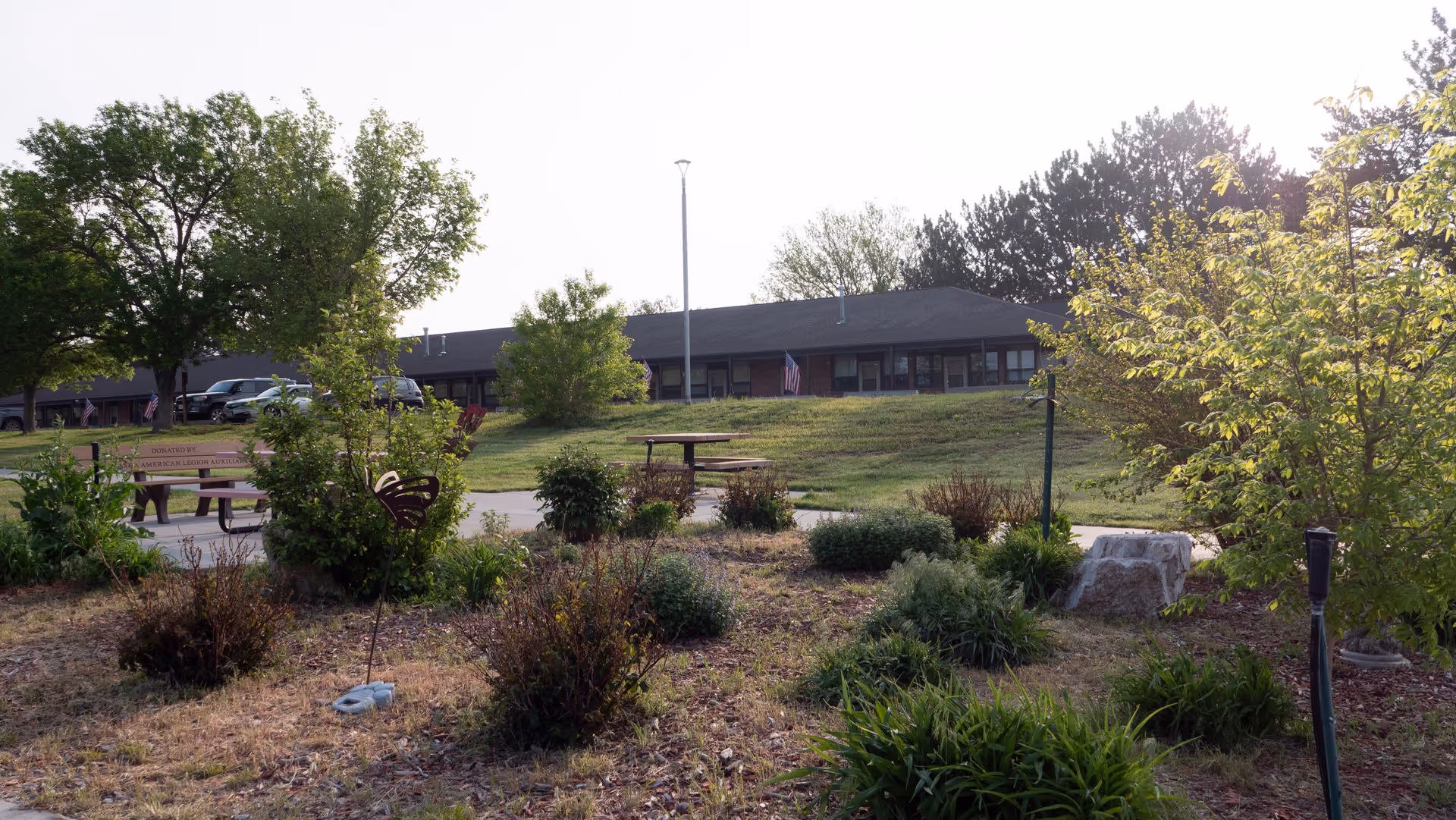 Exterior view of a single-story building set behind a landscaped garden with shrubs, picnic tables, benches, a flagpole, and trees.